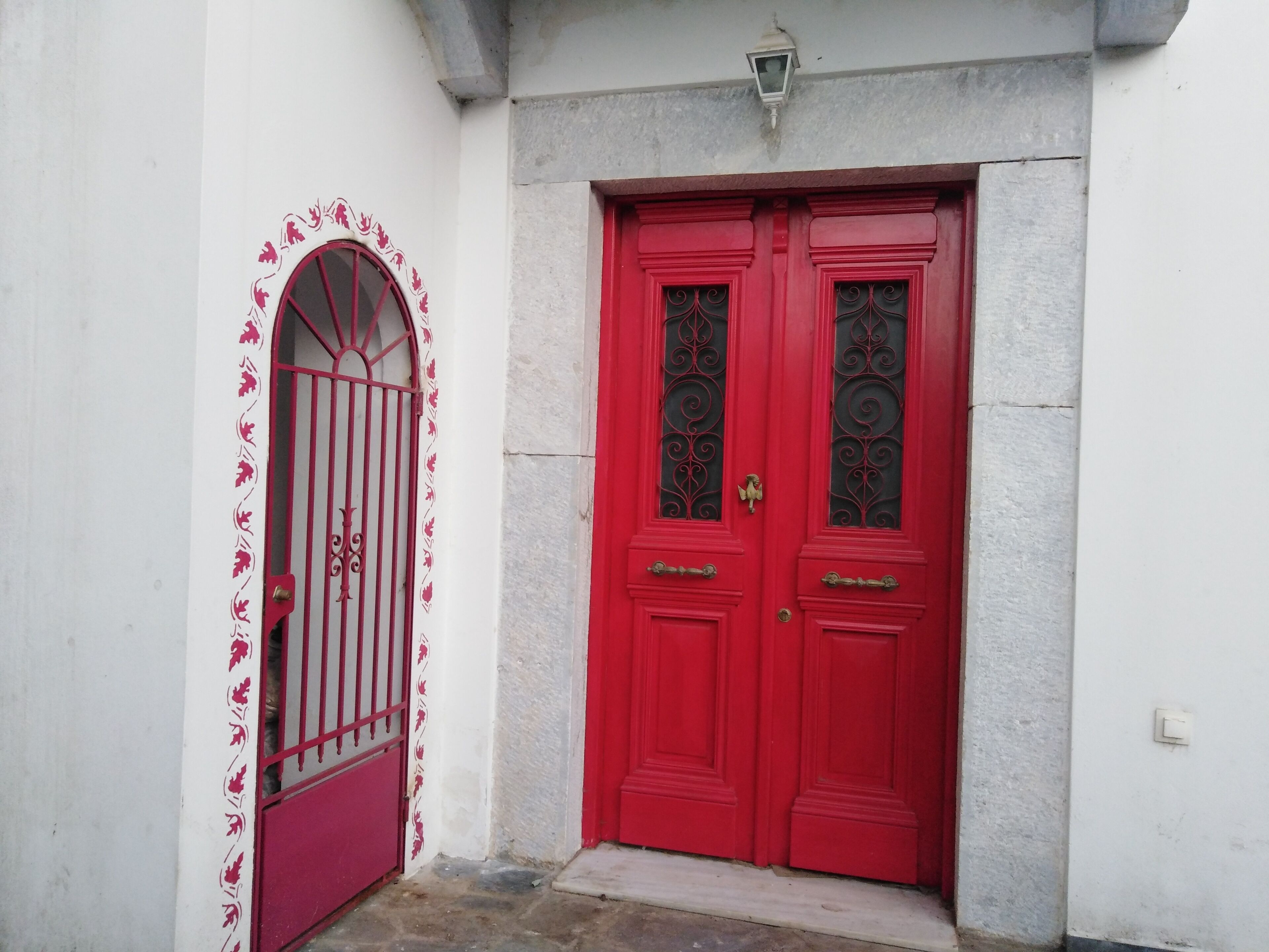Eine rote Doppeltür mit verzierten Glasfeldern und einem Metallgitter, vor einer weißen Wand mit einer kleinen Leuchte darüber.