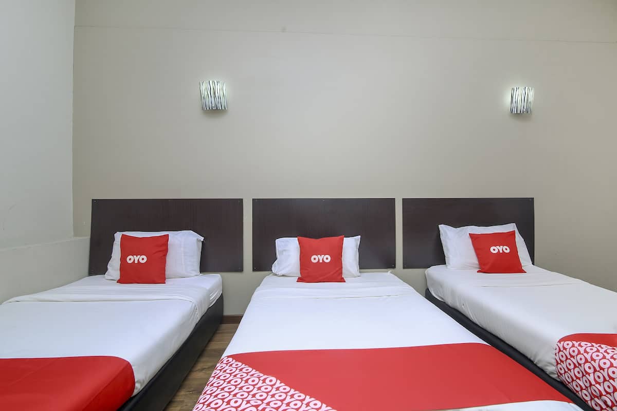 Three single beds with white bedding and red accents, including pillows and a blanket. Each bed has a dark headboard and is positioned against a plain wall with two wall-mounted light fixtures.
