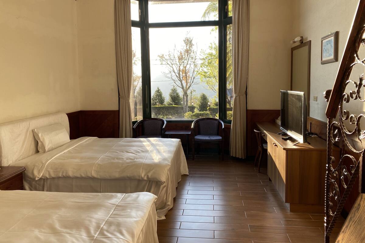 Una habitación de hotel con dos camas, un escritorio con televisor, una silla y un ventanal con cortinas.