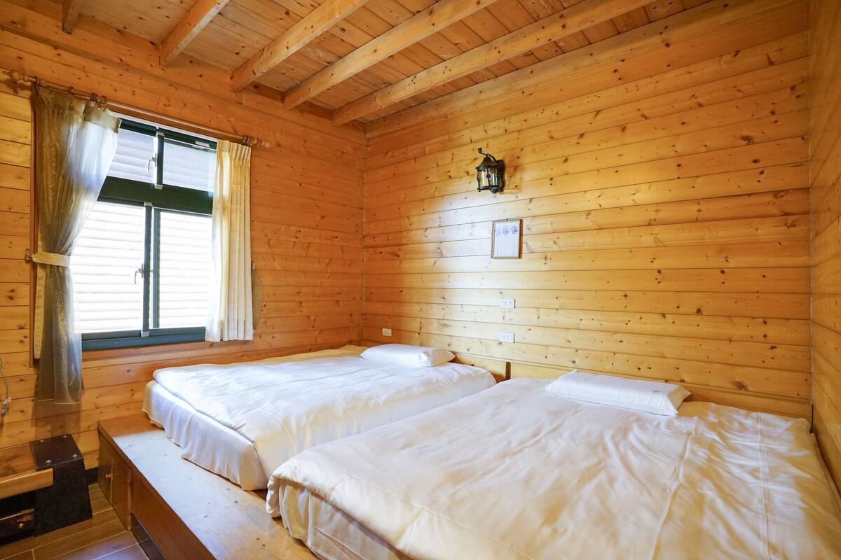 Una cabaña con dos camas de madera, una ventana con cortinas, un farol en la pared y un pequeño marco con un cuadro.