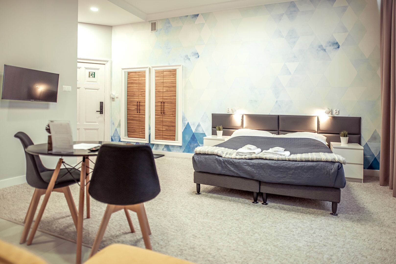 A hotel room with a bed, bedside tables, a desk with chairs, and a TV.