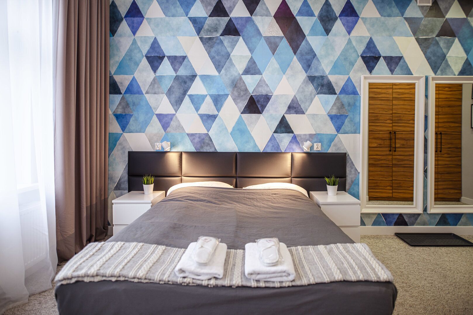 A modern hotel room with a geometric-patterned wall, a large bed with a dark headboard, two bedside tables with lamps, and a wooden wardrobe.