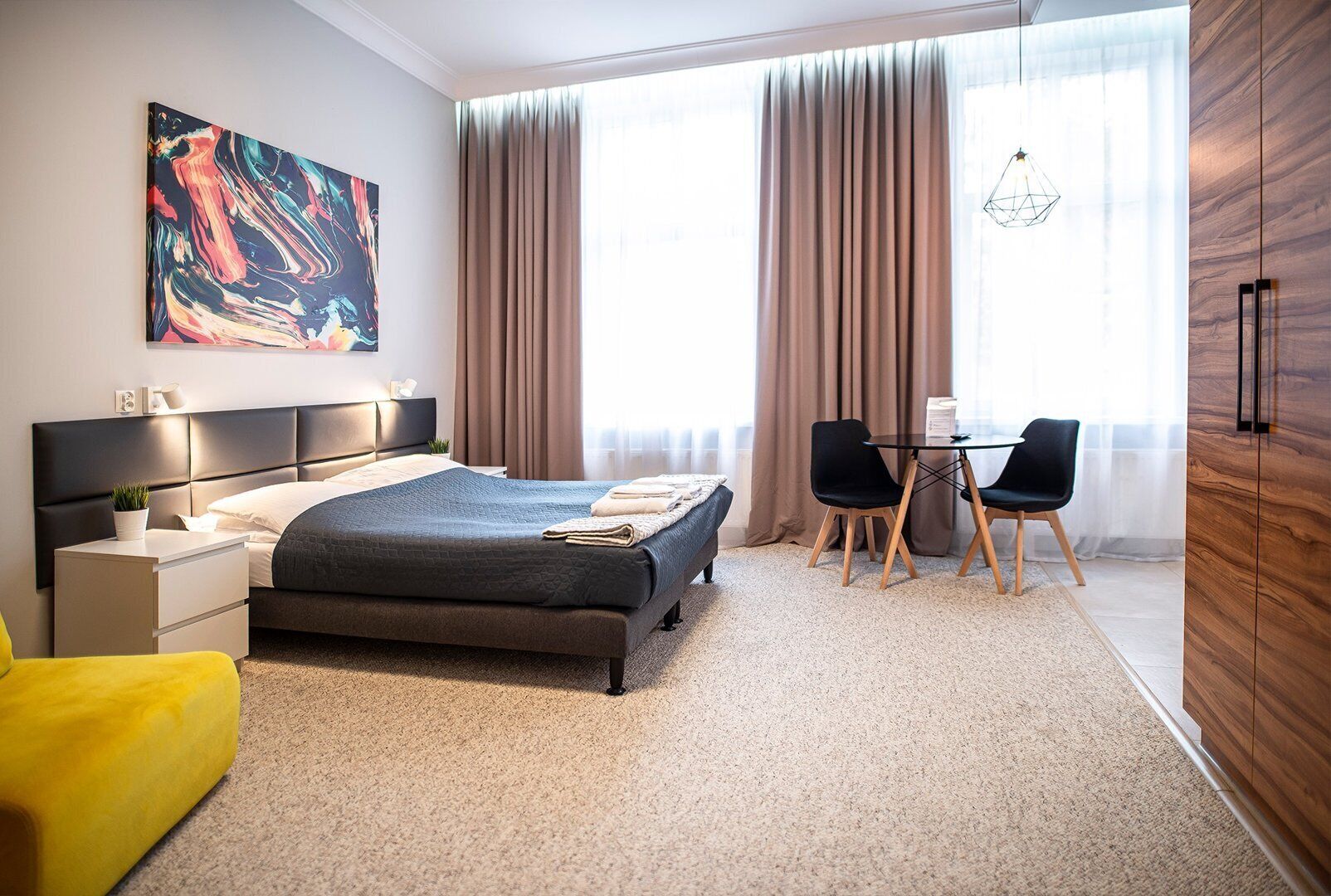 Ein modernes Hotelzimmer mit einem großen Bett, einem Schreibtisch mit zwei Stühlen, einem großen Fenster mit Vorhängen und einem farbenfrohen abstrakten Gemälde an der Wand.