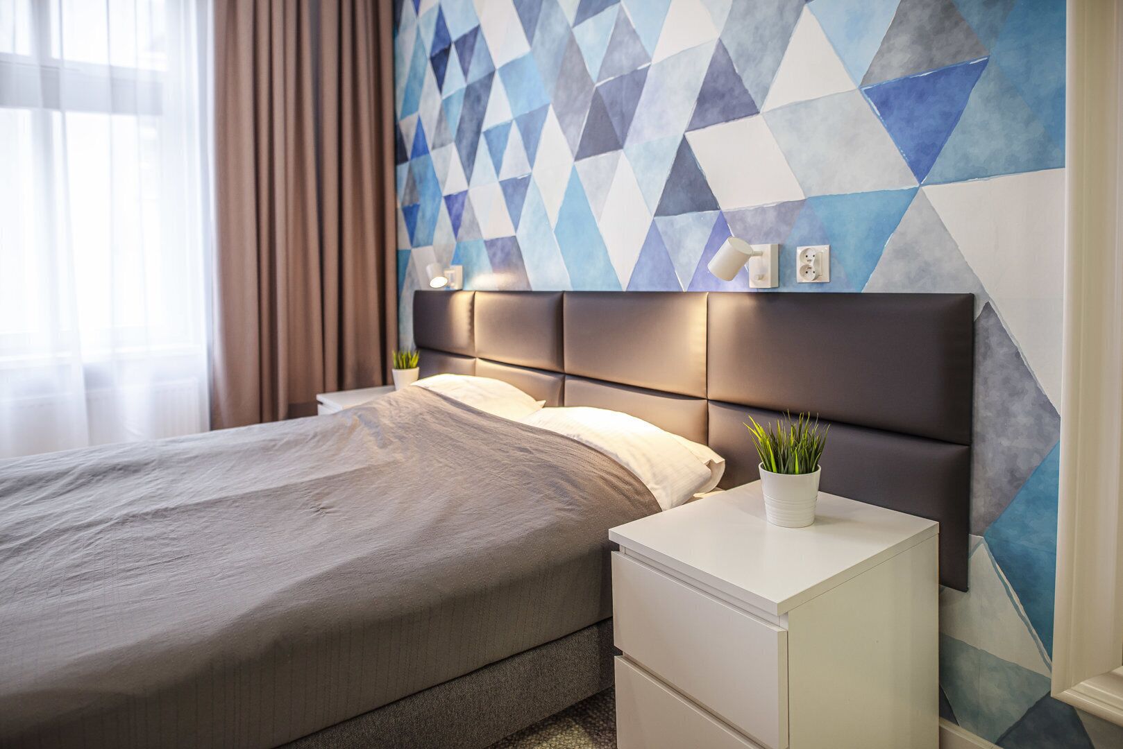 A modern hotel room with a geometric-patterned wall, a bed with a grey bedspread, a white bedside table with drawers, and a potted plant on the table.