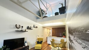 A modern living room with a flat-screen TV, sofa, and yellow armchairs. A dining area with a glass table and chairs is visible in the background. - RWY Suite48 (Plymouth)
