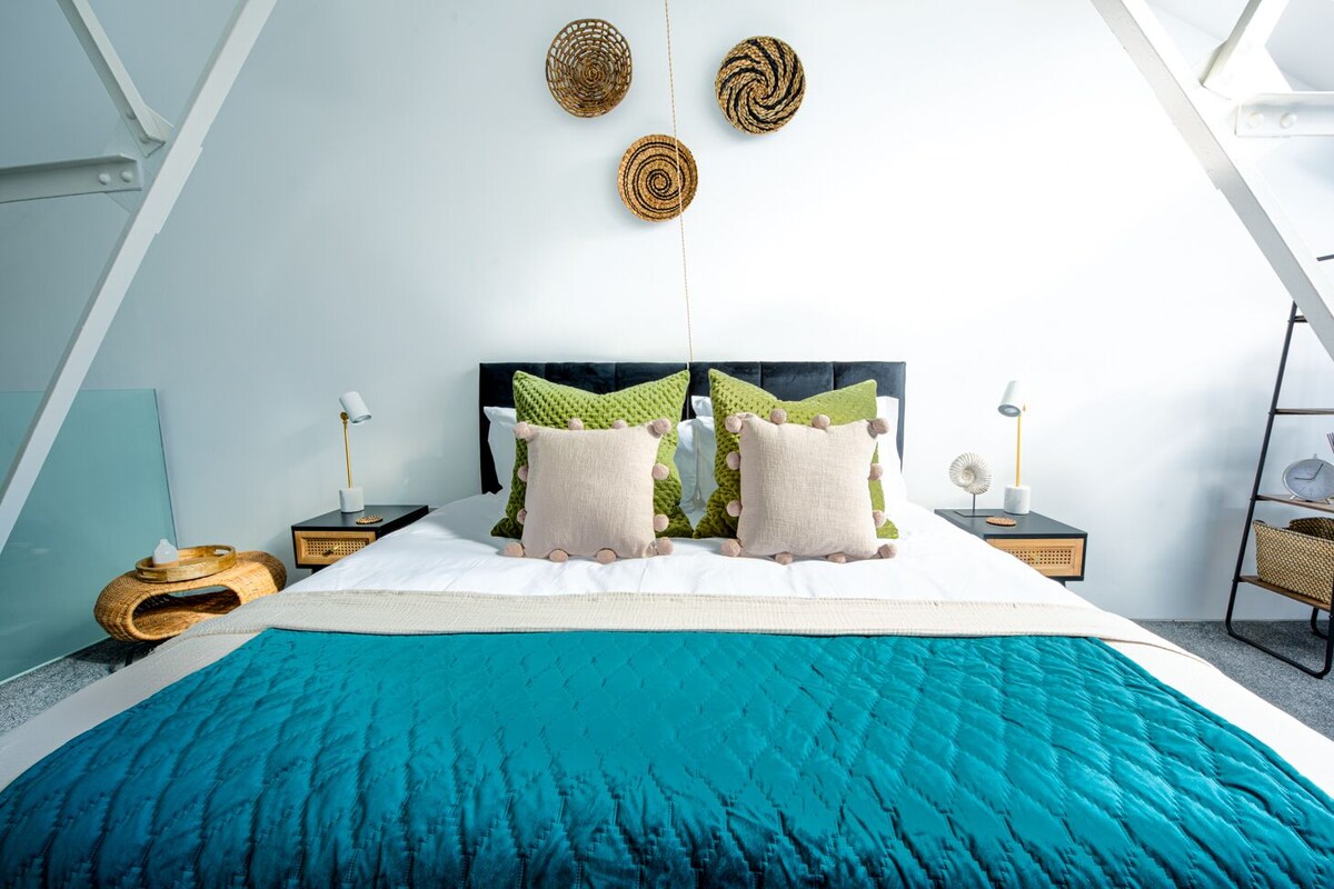 Ein modernes Schlafzimmer mit einem großen Bett, einem schwarzen Kopfteil und einer türkisblauen Bettdecke. Es gibt zwei Nachttische mit Lampen und eine kleine Ablage mit einer Uhr.