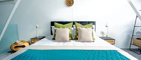 Une chambre à coucher moderne avec un grand lit, une tête de lit noire et une couvre-lit turquoise. On y trouve deux tables de chevet avec des lampes et une petite étagère avec une horloge.