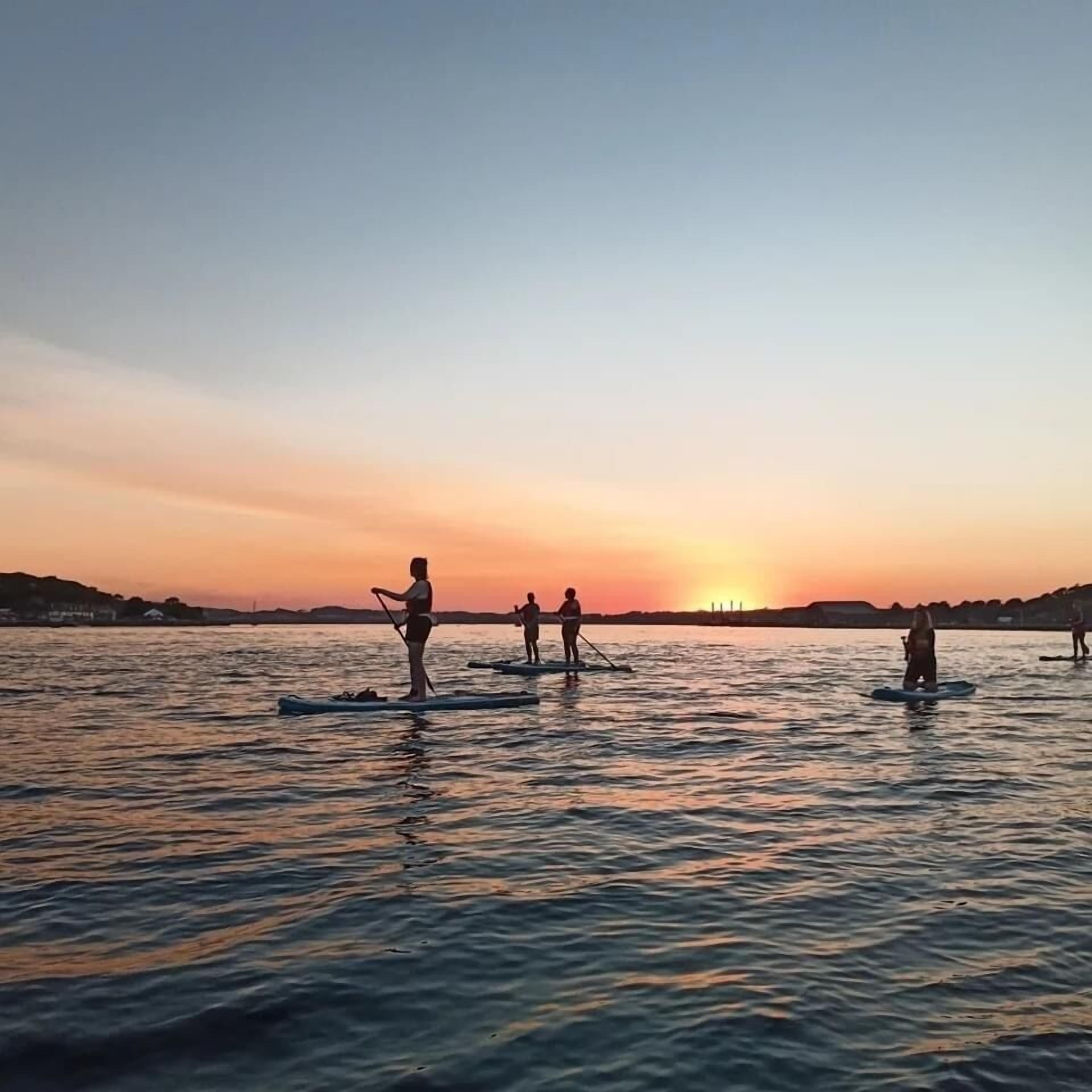 A sunset over a calm body of water with people paddleboarding.