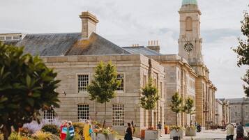 A historic building with a clock tower, a paved courtyard, and a row of trees.