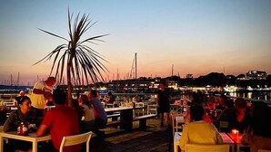 A waterfront dining area with tables and chairs, a palm tree, and boats in the background. - RWY Suite48 (Plymouth)