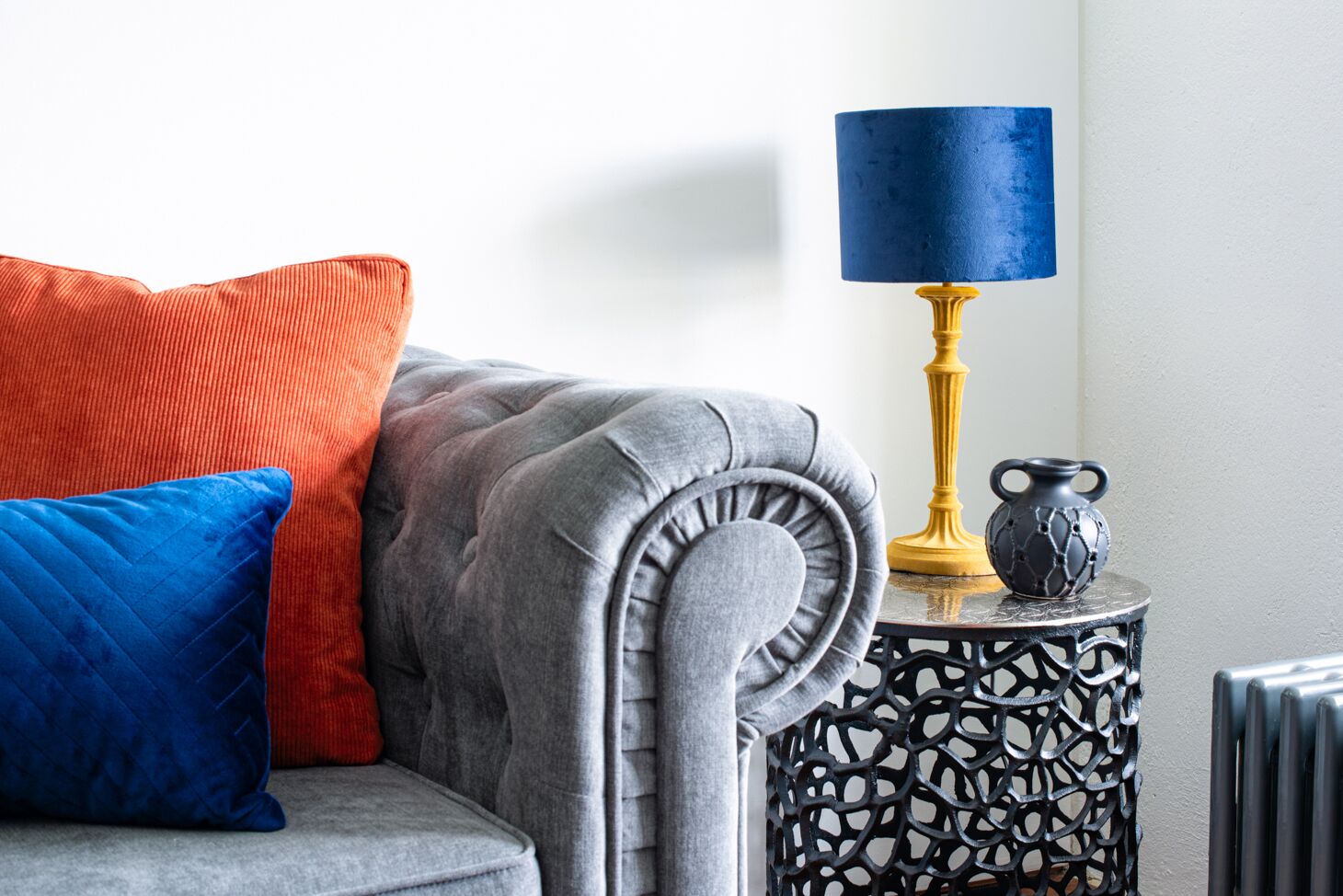 A grey upholstered sofa with a tufted backrest, a blue and red cushion, and a black and gold side table with a blue lampshade.