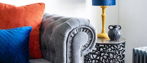 A grey upholstered sofa with a tufted backrest, a blue and red cushion, and a black and gold side table with a blue lampshade.