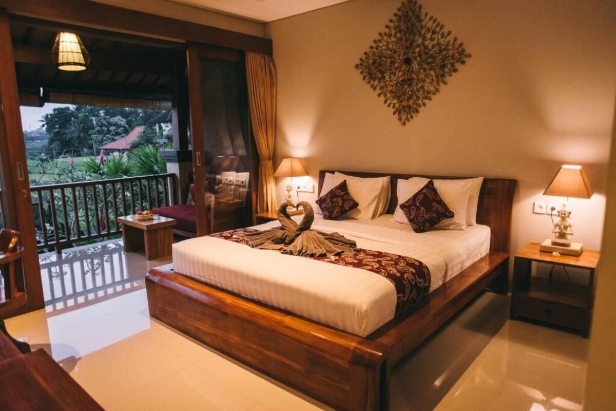 A spacious bedroom with a large bed, two bedside tables, a decorative wall piece, and a balcony with a view of greenery.
