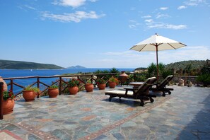 An outdoor patio with stone flooring, potted plants, and lounge chairs with an umbrella. - Villa Elena (Alonissos)