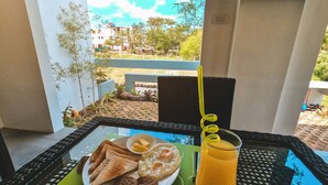 A breakfast table with toast, eggs, sausages, and a glass of orange juice on a balcony overlooking a garden and buildings. - Rold and Roub Home Suites by Cocotel (Puerto Galera)