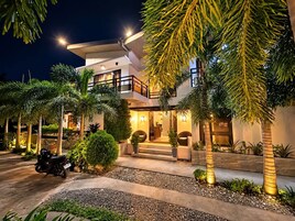 A modern two-story building with a balcony, surrounded by palm trees and a well-lit entrance.