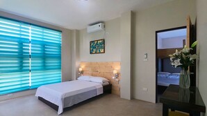 A hotel room with a bed, a desk with a chair, a window with blinds, a wall-mounted air conditioner, and a painting. - Rold and Roub Home Suites by Cocotel (Puerto Galera)