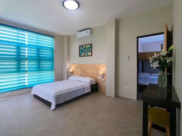 A hotel room with a bed, a desk with a chair, a window with blinds, a wall-mounted air conditioner, and a painting.