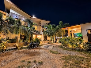 A modern building with a well-lit entrance, a paved driveway, and several motorcycles parked outside. - Rold and Roub Home Suites by Cocotel (Puerto Galera)