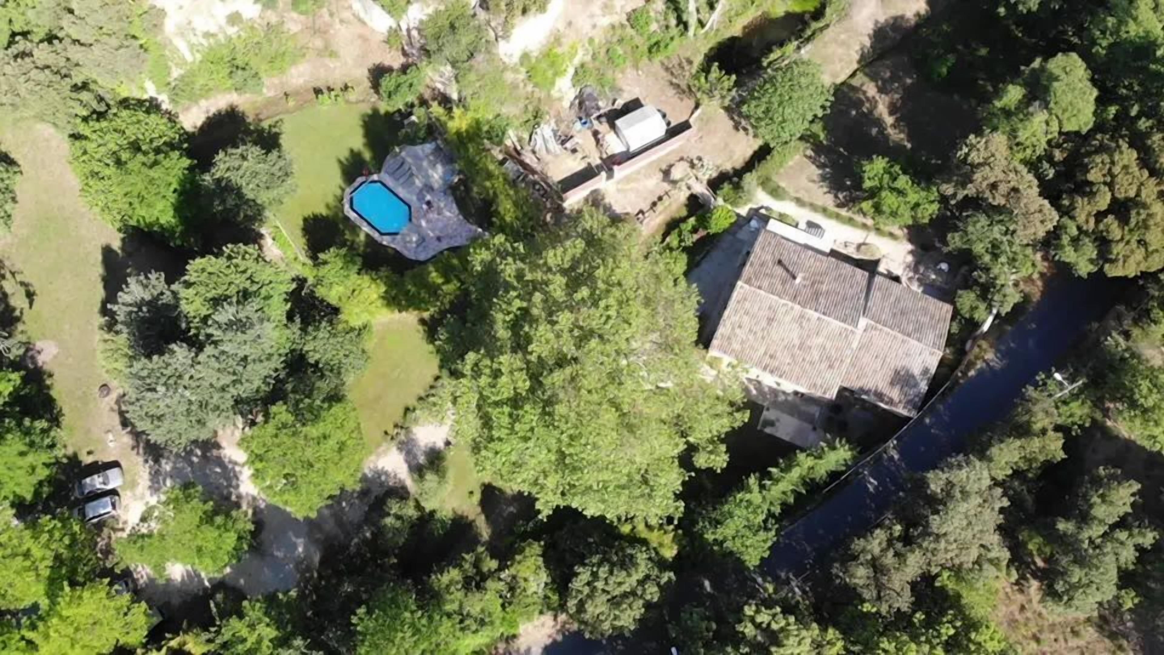 An aerial view of a residential area with a swimming pool, a house with a sloped roof, and dense surrounding vegetation.