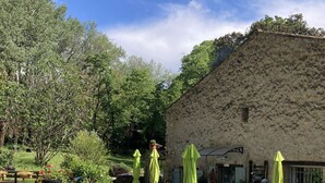 Ein Sitzbereich im Freien mit Tischen und Stühlen, umgeben von Grünpflanzen und einem Steinhaus.