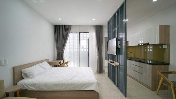 A modern hotel room with a large bed, a TV, a kitchenette, and a view of the city through the window.