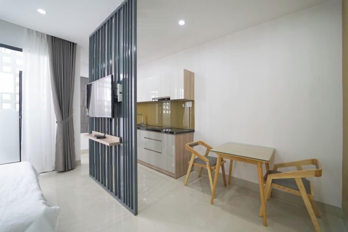 A modern hotel room with a bed, a desk, a chair, a TV, and a kitchenette.
