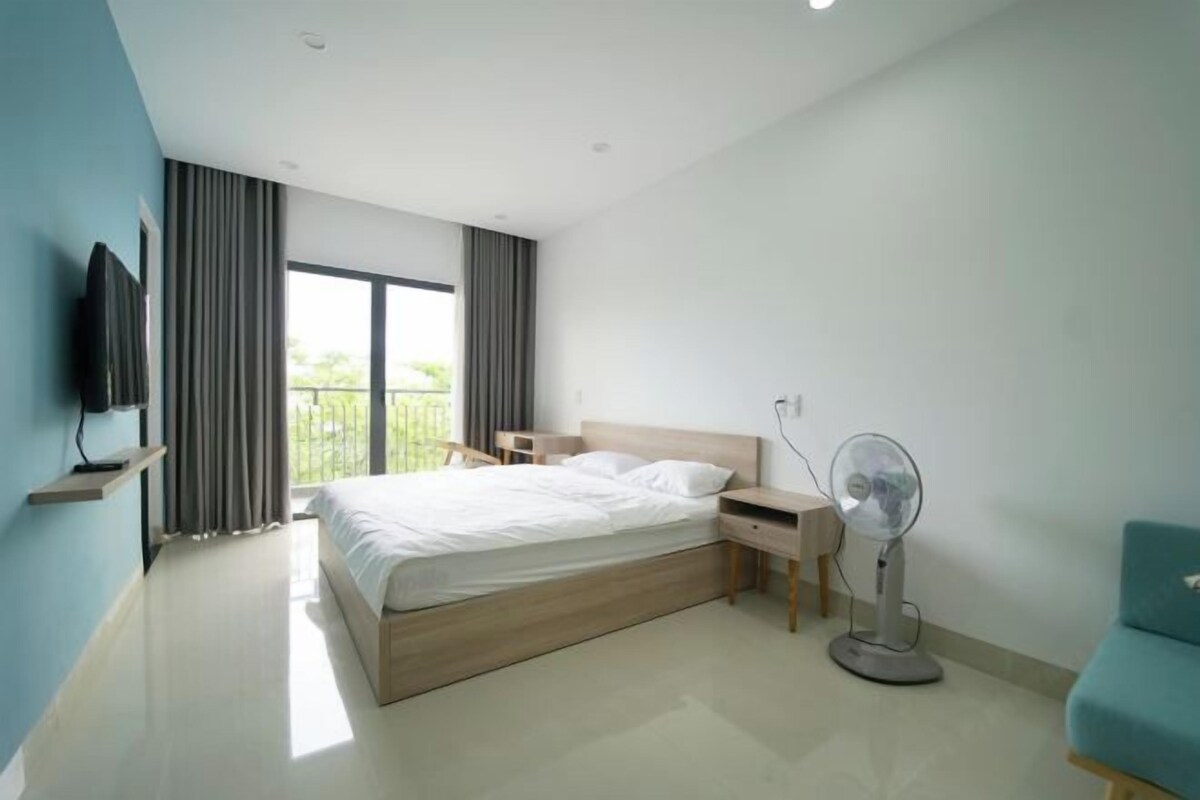 A modern bedroom with a large bed, a bedside table, a standing fan, and a TV mounted on the wall.