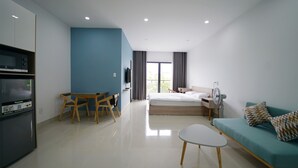 A modern hotel room with a bed, a sofa, a dining table with chairs, a kitchenette, and a large window with curtains.