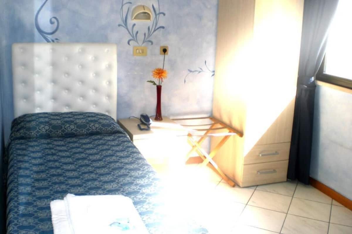 Un dormitorio con una colcha estampada, un cabecero blanco, una mesita de noche con un jarrón de flores y una ventana con cortinas.