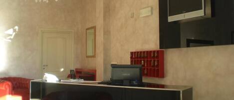 Zona de recepción de un hotel con un mostrador, televisor, candelabro y un área de descanso roja.