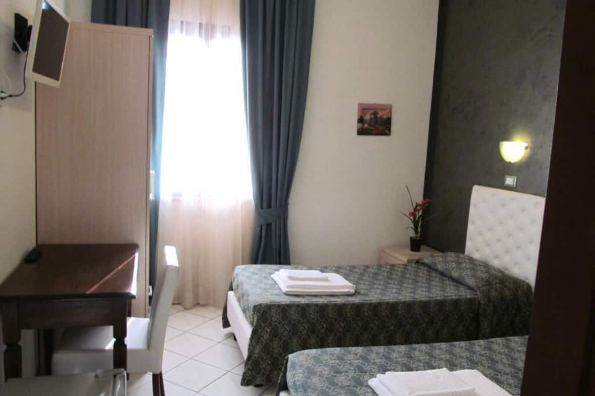 Habitación de hotel con dos camas, un escritorio, una silla, una mesita y un televisor montado en la pared.