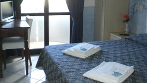 A hotel room with a bed, a desk with a computer, a chair, a nightstand with a vase, and a window with curtains.