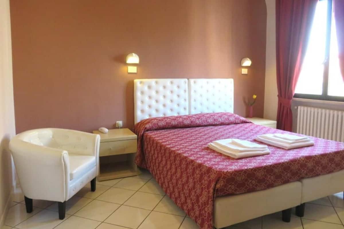 Un dormitorio con cama, una silla, una mesita de noche y una ventana con cortinas.