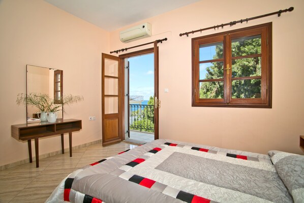 A bedroom with a bed, a wooden desk, a mirror, and a window with a view of trees.