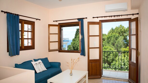 A room with a blue sofa, a white table, and a view of greenery and a balcony. - Hot Tub Cottage Kalymnos (Kalymnos)