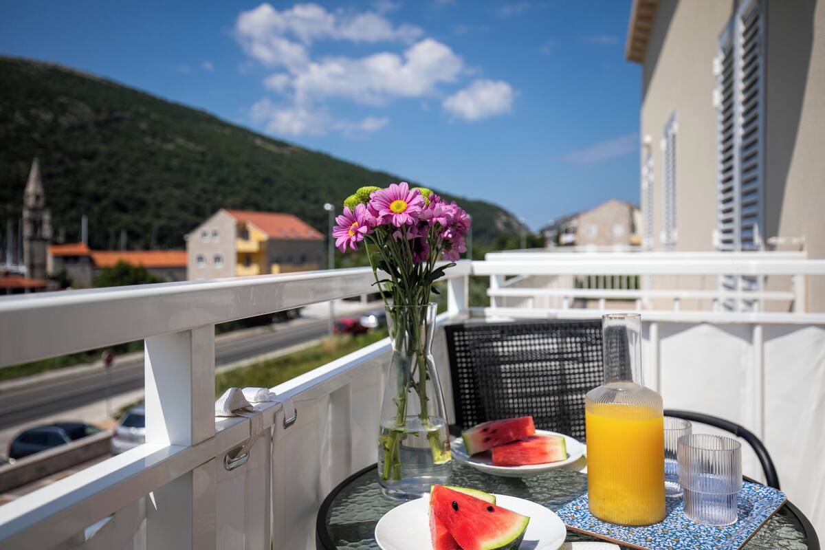 A balcony with a table set for two, featuring a plate of watermelon, a glass of orange juice, and a vase with flowers.