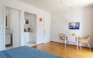A bedroom with a bed, a dining area with chairs and a table, a kitchen with cabinets and a clock, and a hallway.