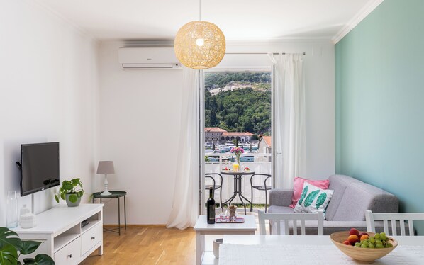 A modern room with a TV, a dining table, a sofa, and a balcony with a view of a marina and buildings.