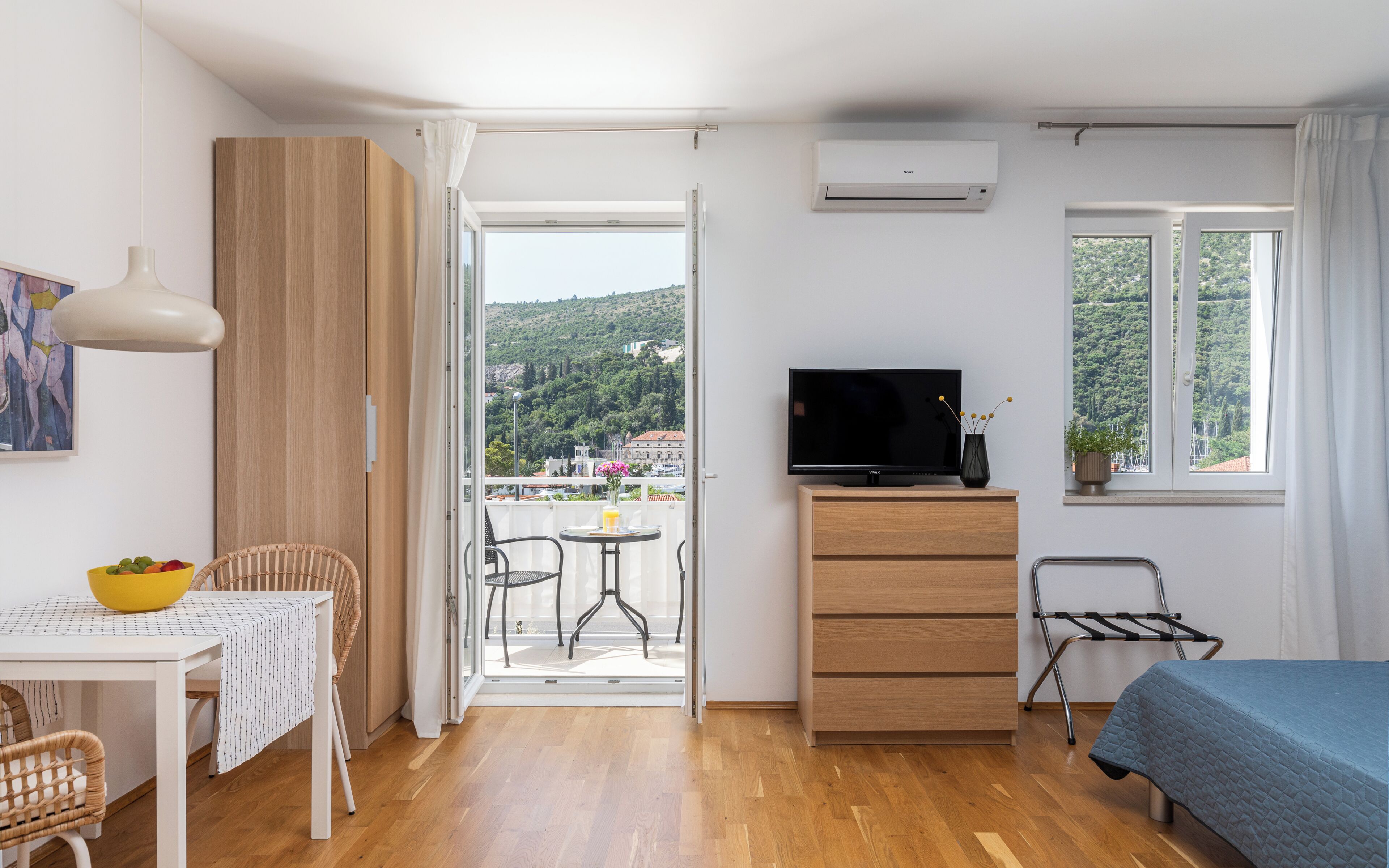 A room with a bed, a TV, a table with chairs, a dining table, a window with a view of a hill, and an air conditioning unit.