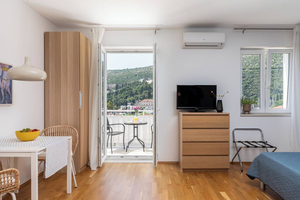 A room with a bed, a TV, a table with chairs, a dining table, a window with a view of a hill, and an air conditioning unit.