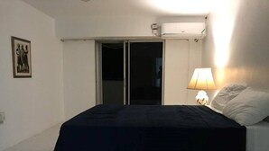 Makuuhuoneessa on sänky, yöpöytä lampilla, ikkuna verhoilla ja ilmastointilaite.