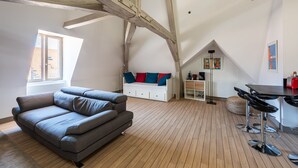 Ein modernes Loft-Apartment mit freiliegenden Holzbalken, einem grauen Sofa, einem Essbereich mit Barhocker und einem Schlafbett im oberen Bereich mit bunten Kissen.