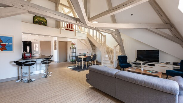 Ein geräumiger Dachboden mit hoher Decke, Holzbalken, einer Küchenzeile mit Barhocker, einem Esstisch und einem Wohnbereich mit Sofa und Fernseher.