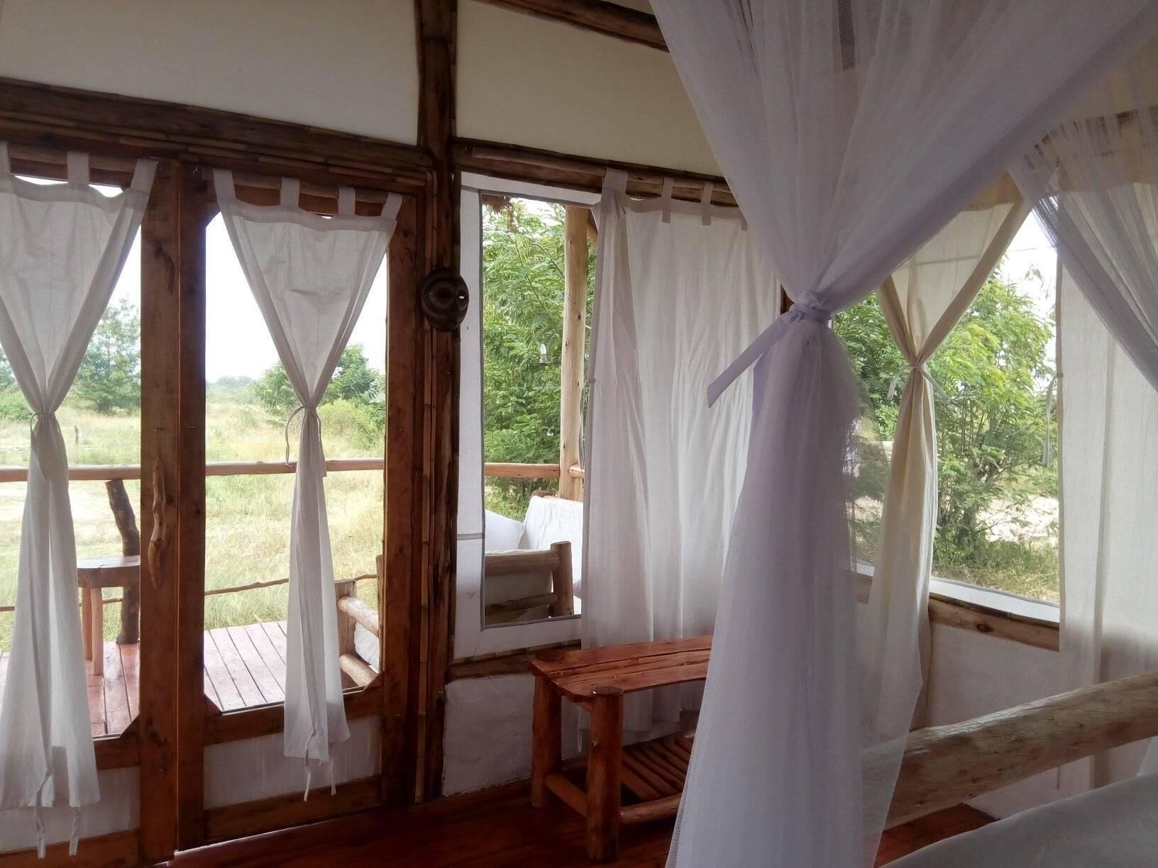 A rustic wooden room with a wooden bench and a table, large windows with white curtains, and a view of greenery outside.