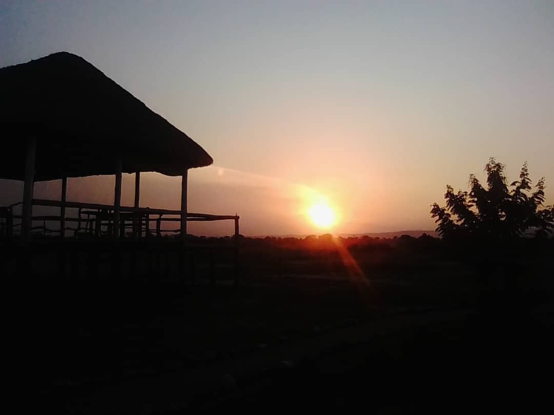 A structure with a roof, possibly a pavilion or shelter, in a natural setting with a sunset in the background.