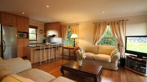 A modern living room with a sofa, coffee table, and TV. A kitchen with wooden cabinets and a refrigerator is visible in the background. - Lavender Cottage Upstairs 2 Bedrooms,2 Bathrooms with Magnificent mountain views (Cape Town)