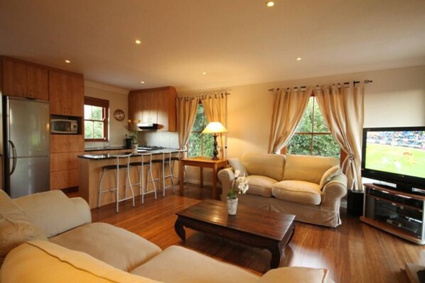 A modern living room with a sofa, coffee table, and TV. A kitchen with wooden cabinets and a refrigerator is visible in the background. - Lavender Cottage Upstairs 2 Bedrooms,2 Bathrooms with Magnificent mountain views (Cape Town)