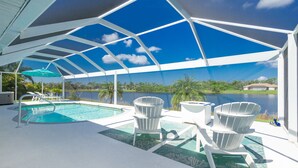 A covered outdoor area with a pool, two Adirondack chairs, and a view of a lake.