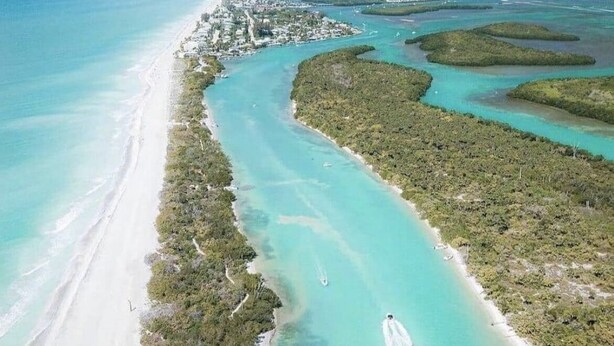 An aerial view of a coastal area with a long stretch of beach, clear turquoise waters, and a dense green shoreline.
